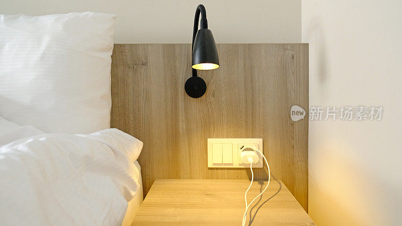 Close-up fragment of bedroom with empty bedside table, turn on reading lamp and a socket in modern interior​ design home or hotel. Soft pillow and blanket, stylish comfortable furniture.